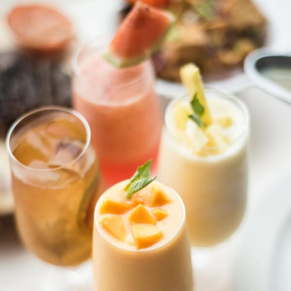 Four vibrant Thai drinks: iced tea, watermelon, pineapple, and mango smoothies, garnished with fresh fruit.